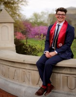 Logan smiles while wearing a suit and sitting on a sign that says Indiana University